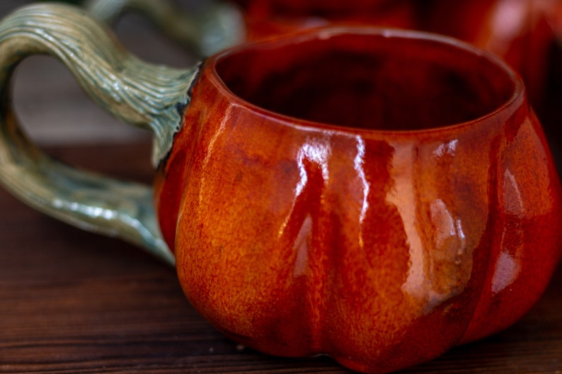 May include: A ceramic pumpkin-shaped mug with a green handle. The mug is glazed in a deep orange color with a glossy finish.