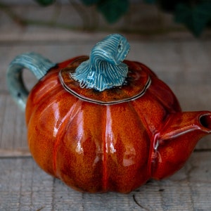 Op de afbeelding: Een keramische theepot in de vorm van een pompoen met een blauw en groen deksel. De theepot is geglazuurd in een glanzende oranje kleur.