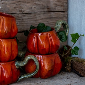 May include: A set of four orange ceramic mugs shaped like pumpkins. The mugs have green handles and are stacked on top of each other. The mugs are sitting on a wooden surface.