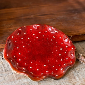 May include: A small, round, ceramic dish with a glossy, deep red glaze and a textured, wavy edge. The surface is decorated with numerous small, white, raised dots scattered across the interior, creating a speckled appearance on the red base.