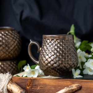 May include: Bronze-colored ceramic mug with a textured, scale-like pattern. The mug has a curved handle and sits on a wooden tray with white flowers. A matching decorative sphere is in the background. The mug is designed for hot beverages.