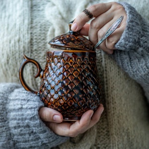 Könnte beinhalten: Eine braune Keramiktasse mit einem strukturierten, schuppigen Design. Die Tasse hat einen Deckel und einen Griff. Die Tasse wird von einer Person gehalten, die einen Strickpullover trägt.