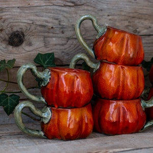 May include: A set of four ceramic pumpkin mugs with green handles. The mugs are stacked on top of each other, with the largest mug at the bottom and the smallest mug at the top. The mugs are all glazed in a glossy orange color.
