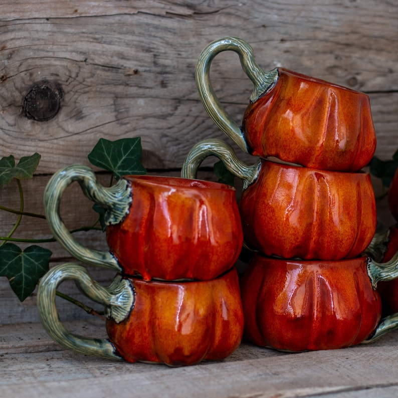 May include: A set of four ceramic pumpkin mugs with green handles. The mugs are stacked on top of each other, with the largest mug at the bottom and the smallest mug at the top. The mugs are all glazed in a glossy orange color.