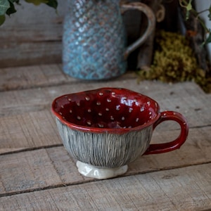 Könnte beinhalten: Eine keramische, pilzförmige Tasse mit roter Innenseite, die mit weißen Punkten versehen ist. Die Außenseite ist grau mit vertikalen Linien und hat einen roten Henkel. Die Tasse steht auf einem weißen, wolkenartigen Sockel. Im Hintergrund befindet sich ein türkisfarbener Krug.