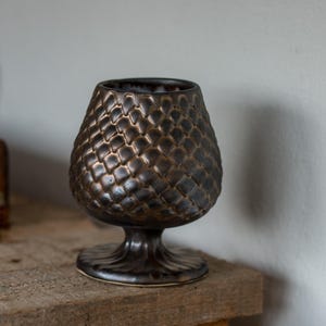 Peut inclure: Un gobelet en céramique brun foncé présentant un motif texturé en forme d'écailles sur son bol arrondi. Le récipient repose sur une base courte et évasée, avec un bord intérieur lisse et une finition émaillée aux tons métalliques profonds.