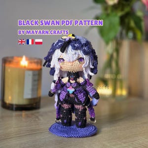 May include: Crocheted doll of a character with white and purple hair, wearing a purple and black outfit. The doll is on a purple base. Text on the image reads "BLACK SWAN PDF PATTERN BY MAYARN.CRAFTS". A lit candle is in the background.