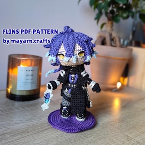 Puede incluir: Muñeca de ganchillo hecha a mano con cabello azul y morado, vestida con un atuendo negro con detalles plateados. La muñeca está sobre una base de ganchillo morada. Se ve el texto "FLINS PDF PATTERN by mayarn.crafts".