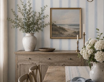May include: A room featuring wallpaper with vertical blue and white stripes, a wooden sideboard, a framed coastal landscape painting, a ceramic vase with greenery, a wooden table with a striped runner, and the text KITTEN LANE WALLPAPER & TEXTILES.