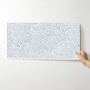 May include: A roll of wallpaper featuring a repeating pattern of stylized, light blue coral branches on a white background. The wallpaper is being held up against a white wall, showcasing the design.