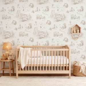 May include: A light wood crib with a white mattress and a cream-colored blanket, positioned against wallpaper featuring a repeating brown line-art pattern of characters, including a bear and a balloon, in a nursery setting with a lamp and basket.