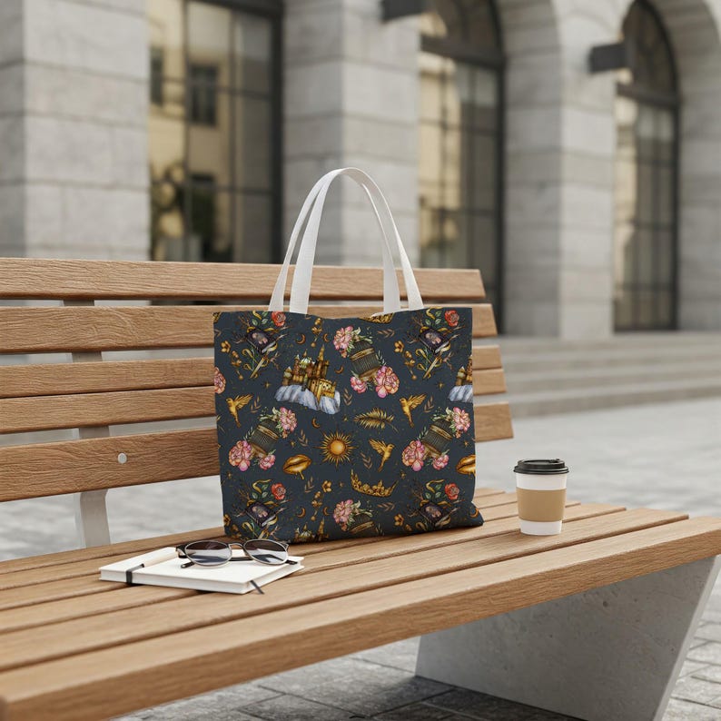 May include: A dark blue tote bag with white handles, featuring a repeating pattern of flowers, books, suns, and hummingbirds. The bag is sitting on a wooden bench with a notebook, glasses, and a coffee cup.