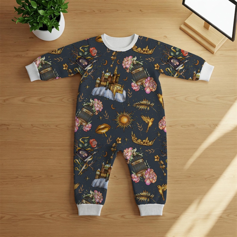 May include: A dark blue baby onesie with a white collar and cuffs. The onesie is covered in a whimsical pattern of castles, crowns, flowers, birds, and suns. The design is in shades of pink, gold, and white.
