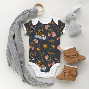 May include: A dark blue baby onesie with a floral and celestial pattern, featuring flowers, suns, and birds. The image also includes brown fringed baby boots, a gray bunny toy, and a wooden ring.