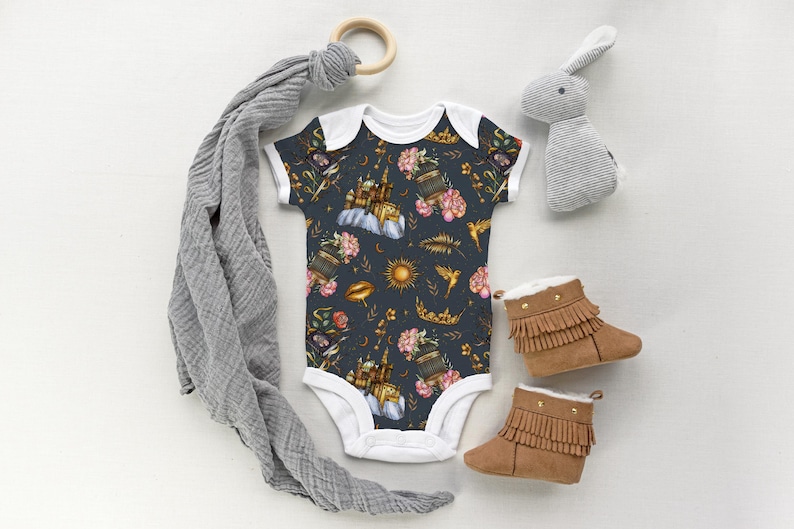 May include: A dark blue baby onesie with a floral and celestial pattern, featuring flowers, suns, and birds. The image also includes brown fringed baby boots, a gray bunny toy, and a wooden ring.