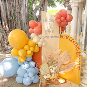 Könnte beinhalten: Eine Babyparty-Dekoration mit einem gelben und orangen Sonnenhintergrund, einem Schild mit der Aufschrift "Willkommen zur Babyparty von Julie" und einem Weidenkinderwagen mit Pampasgras.