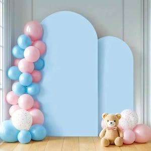 Puede incluir: Un telón de fondo azul claro con dos recortes en forma de arco está decorado con globos rosas y azules. Un oso de peluche con una pajarita rosa se sienta frente al telón de fondo, con más globos rosas y blancos.