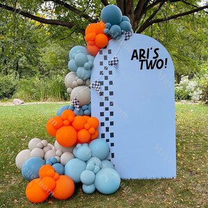 May include: A blue and white archway backdrop with a chequered pattern and the text "Ari's Two!"  The archway is decorated with a garland of orange, blue, and grey balloons.