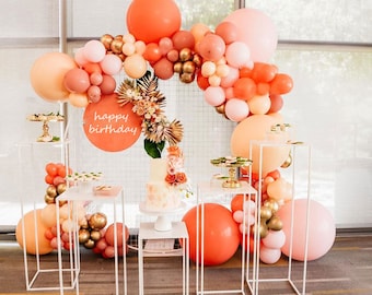 May include: A birthday party decoration with a balloon arch made of peach, pink, and gold balloons. The arch is decorated with greenery and a sign that says "happy birthday". There are also white cake stands with desserts on them.