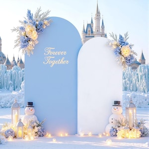 May include: A light blue and white wedding backdrop with the text "Forever Together" in gold script. The backdrop is decorated with floral arrangements, snowmen, and lanterns. A castle is visible in the background.