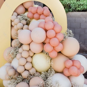 May include: A gold archway decorated with a variety of balloons in shades of beige, peach, and white. The balloons are arranged in a cascading pattern, with the largest balloons at the bottom and the smallest balloons at the top. The archway is decorated with small white flowers.