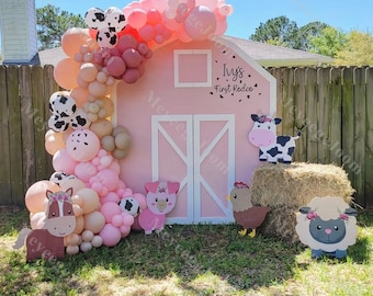 May include: A pink barn backdrop with a white door and a sign that reads "Ivy's First Rodeo". The backdrop is decorated with a balloon arch in shades of pink, beige, and white. There are also farm animal cutouts, including a cow, pig, chicken, and sheep.