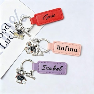 May include: Three personalized graduation keychains. Each keychain has a different colored leather tag with a name: Susie, Rafina, and Isabel. Each keychain has a silver charm with a graduation cap, diploma, and the year 2026.