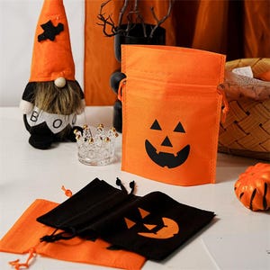 Op de afbeelding: Oranje Halloween-snoerzakken met zwarte pompoengezichten. Links staat een Halloween-kabouter met een oranje hoed en een zwart pak. Een kleine decoratieve kroon en een miniatuurpompoen zijn ook zichtbaar.