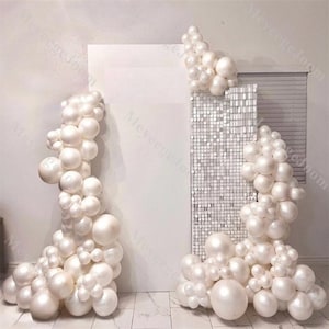 May include: White balloon arch backdrop with a white sequin wall in the center. The balloons are arranged in a cascading design, creating a festive and elegant atmosphere.