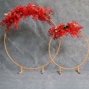 May include: Two gold metal hoops with stands, decorated with red artificial flowers and greenery. The hoops are arranged in a nested configuration, with the larger hoop behind the smaller hoop.