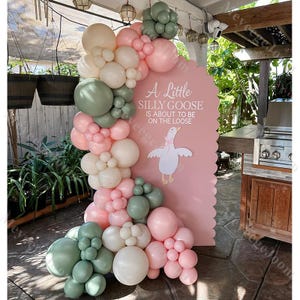 Puede incluir: Un letrero rosa con borde festoneado con el texto "A Little Silly Goose Is About To Be On The Loose" y una ilustración de un ganso de dibujos animados. Un arco de globos en tonos rosa, blanco y verde enmarca el letrero, creando una decoración festiva.