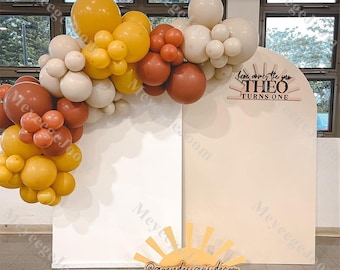 May include: A beige and white backdrop with a wooden sign that reads "Here comes the sun THEO TURNS ONE". A balloon arch made of cream, yellow, and orange balloons is in front of the backdrop. A wooden sun cutout with the text "@greydesignsdecor" is in the foreground.
