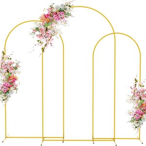 May include: A gold metal wedding arch with three connected arched frames. The arch is decorated with pink, white, and green flowers. The arch is designed for outdoor or indoor events.