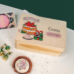 Könnte beinhalten: Eine Holzkiste mit einem Geburtstagskuchen-Design, dem Namen "Emma" und dem Datum "03.07.1987" aufgedruckt. Ein kleiner Kuchen mit Beeren und einer Kerze, eine Karte und verpackte Süßigkeiten sind ebenfalls zu sehen.