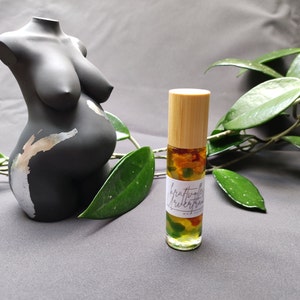 May include: A black, pregnant torso statue with silver accents stands next to a roll-on essential oil bottle. The bottle has a bamboo cap and a label with text. Green leaves and a gray background complete the scene.