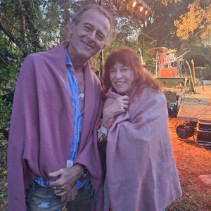 May include: Two people are wrapped in pink shawls, smiling at the camera. The man wears a blue shirt and ripped jeans. The woman wears a patterned top and jeans. A stage with musical instruments is visible in the background.