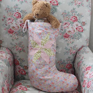 Puede incluir: Un calcetín navideño de tela con un estampado floral azul claro y un diseño central de patchwork en forma de estrella en tonos rosa suave y verde salvia. Un pequeño oso de peluche marrón está dentro del calcetín, que tiene una pequeña asa.