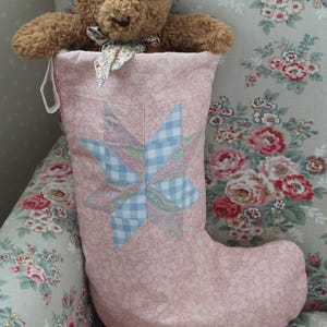 Puede incluir: Un calcetín navideño de tela rosa claro con un diseño central de estrella de patchwork en cuadros azules y motivos florales. Un oso de peluche marrón con un pequeño pañuelo floral en el cuello está metido en la abertura superior.