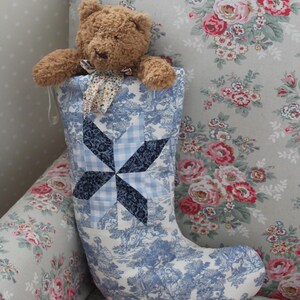 May include: A brown plush teddy bear sits inside a fabric Christmas stocking. The stocking features a blue and white toile de jouy pattern with a central navy and light blue gingham star quilt block design and a small floral ribbon at the neck.