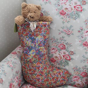 Könnte beinhalten: Ein gesteppter Weihnachtsstrumpf aus Stoff mit einem dichten, mehrfarbigen Blumenmuster in Rot-, Blau-, Gelb- und Grüntönen. Ein kleiner, hellbrauner Plüschteddybär mit einem winzigen gemusterten Halstuch sitzt in der Öffnung des Strumpfes.