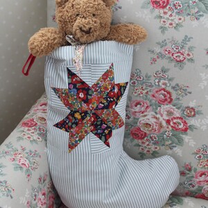 May include: A light blue and white pinstriped fabric stocking featuring a large, multi-colored floral star appliqué on the front. A small, brown, fuzzy teddy bear with a floral patterned neck ribbon is tucked inside the top of the stocking.
