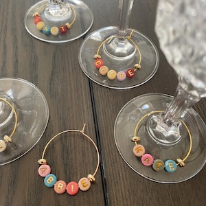 May include: Wine glass charms with gold hoops and colourful letter beads spelling out names. The charms are attached to the base of the wine glasses. Names include Annette, Clare, Abbie, and Kate.