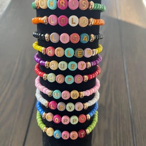 May include: A collection of beaded bracelets in various colours, each spelling out a name in gold lettering. The bracelets include names like "CHRIS", "LOLA", and "LISA". The beads are arranged on elastic, creating a vibrant and personalized accessory.