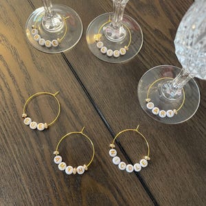 May include: A set of gold-toned metal wine glass charms featuring white circular beads with gold lettering. The charms spell out the names Bride, Alice, Gary, Sue, Amy, and Nicole, with small gold-coloured spacer beads between the lettered beads.