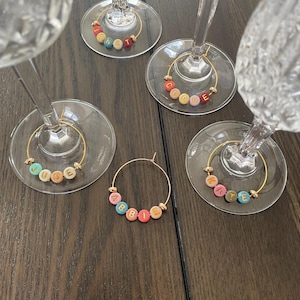 May include: Wine glass charms with gold-coloured hoops and colourful letter beads spelling out names. The charms are attached to the base of wine glasses. Names include: Annette, Clare, June, Abbie, and Tate. The beads are in various colours.