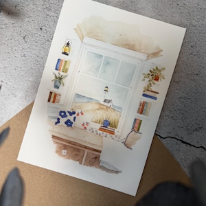 Könnte beinhalten: Aquarelldruck eines Küstenmotivs. Das Kunstwerk zeigt ein Fenster mit Blick auf einen Strand mit einem Leuchtturm. Bücherregale flankieren das Fenster, und ein brauner Schreibtisch befindet sich im Vordergrund. Die Farbpalette umfasst Blau-, Braun- und Weißtöne.