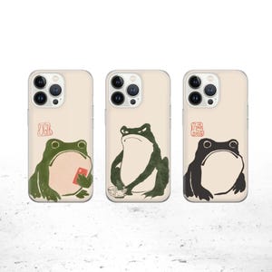 May include: Three iPhone cases featuring different frog illustrations. The cases are clear and display the phone's camera lenses. The frogs are depicted in various poses, one holding a red object, one holding a bowl, and one with its mouth open.