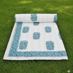 May include: A white and blue floral patterned quilt, partially rolled up, rests on green grass. The quilt features a repeating blue floral design and a blue border with a leaf pattern. The quilt is made of soft material.