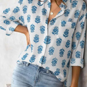 May include: White shirt with a blue floral design. The long-sleeved shirt features a collar, a chest pocket, and rolled-up sleeves. It is worn with light blue denim jeans.