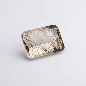 Op de afbeelding: Een rechthoekige edelsteen met een lichtbruine tint en ingewikkelde zwarte naaldachtige insluitsels. De edelsteen heeft een facetgeslepen slijpvorm, die de helderheid en interne patronen laat zien. De steen staat tegen een witte achtergrond.